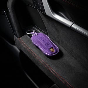 May include: A purple suede key fob cover with white stitching and a silver key ring. The cover features embossed buttons and a Porsche emblem. The key fob is resting on a black, textured surface with red stitching.