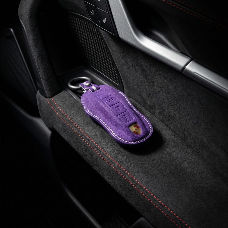 May include: A purple suede key fob cover with white stitching and a silver key ring. The cover features embossed buttons and a Porsche emblem. The key fob is resting on a black, textured surface with red stitching.