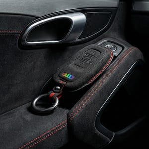 K&ouml;nnte beinhalten: Eine schwarze Wildleder-Autoschl&uuml;sselh&uuml;lle mit einem regenbogenfarbenen Audi-Logo und roten N&auml;hten. Der Schl&uuml;sselanh&auml;nger ist an einem schwarz-roten Schl&uuml;sselband mit einem Metallring befestigt. Der Schl&uuml;sselanh&auml;nger liegt auf einem schwarzen Innenraum mit roten N&auml;hten.