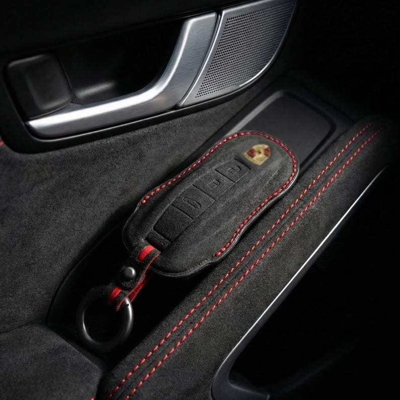 May include: A black leather key fob case with red stitching and a Porsche emblem. The case is resting in a car's interior, next to a door handle and speaker.