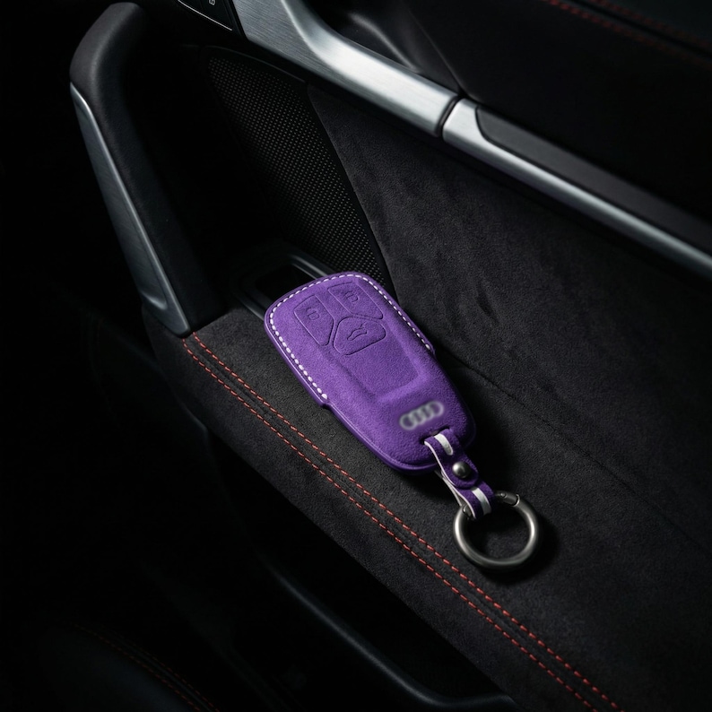 K&ouml;nnte beinhalten: Eine lila Autoschl&uuml;sselh&uuml;lle mit wei&szlig;en N&auml;hten und einem silbernen Schl&uuml;sselring. Die H&uuml;lle hat das Audi-Logo und Schaltfl&auml;chensymbole. Der Schl&uuml;sselanh&auml;nger liegt auf einer schwarzen, strukturierten Oberfl&auml;che mit roten N&auml;hten.
