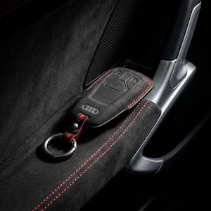 K&ouml;nnte beinhalten: Schwarze Autoschl&uuml;sselh&uuml;lle mit roten N&auml;hten und einem Metallschl&uuml;sselring. Die H&uuml;lle hat das Audi-Logo und Schaltfl&auml;chensymbole. Der Schl&uuml;sselanh&auml;nger liegt auf einer schwarzen, Wildleder-&auml;hnlichen Oberfl&auml;che mit roten N&auml;hten.
