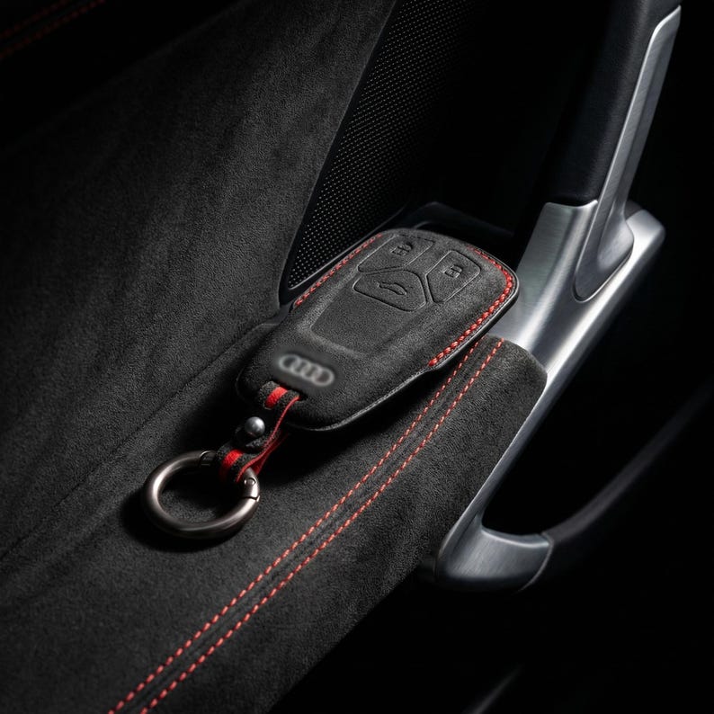 K&ouml;nnte beinhalten: Schwarze Autoschl&uuml;sselh&uuml;lle mit roten N&auml;hten und einem Metallschl&uuml;sselring. Die H&uuml;lle hat das Audi-Logo und Schaltfl&auml;chensymbole. Der Schl&uuml;sselanh&auml;nger liegt auf einer schwarzen, Wildleder-&auml;hnlichen Oberfl&auml;che mit roten N&auml;hten.