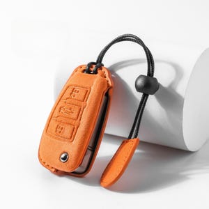 Pode incluir: Capa laranja para chave de carro com s&iacute;mbolos de chave em relevo. A capa tem um cord&atilde;o preto com uma conta preta e uma pequena aba laranja. A capa da chave &eacute; feita de um material semelhante a camur&ccedil;a.