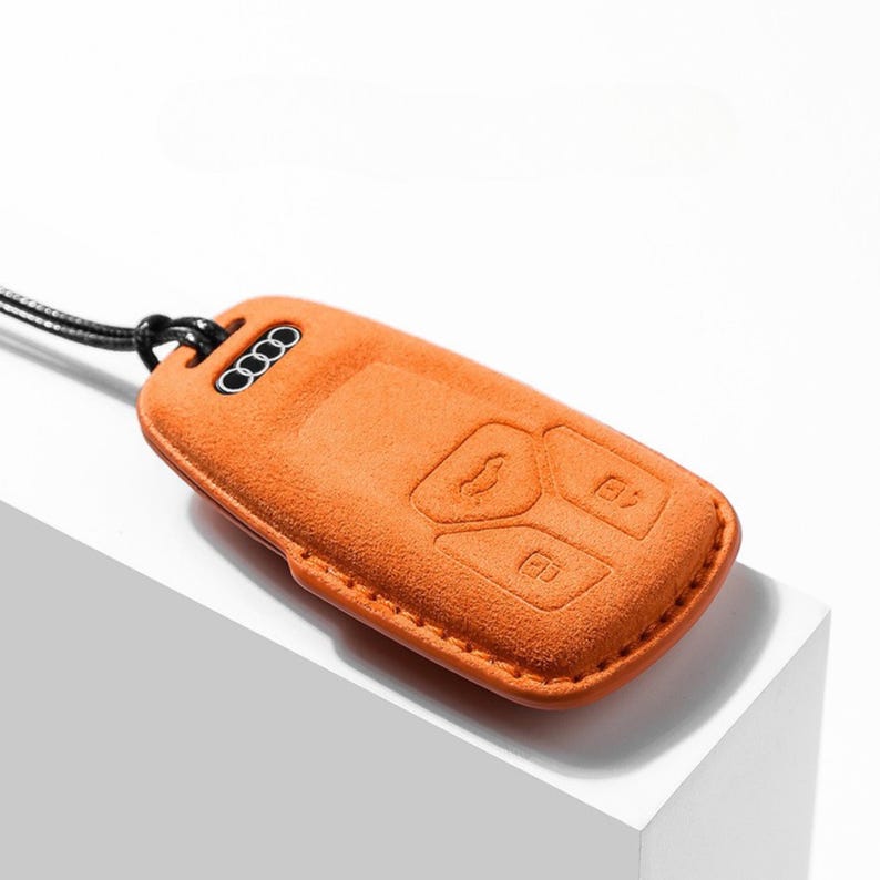 Pode incluir: Capa laranja para chave do carro com bot&otilde;es em relevo e o log&oacute;tipo da Audi. A capa da chave &eacute; feita de um material semelhante a camur&ccedil;a e tem um cord&atilde;o preto. A capa &eacute; retangular e tem uma borda costurada.