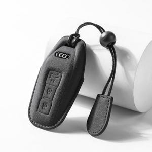 Pode incluir: Uma capa preta para chave de carro com o logotipo da Audi e &iacute;cones de bot&otilde;es. A capa tem um cord&atilde;o com uma conta preta e uma aba de couro. A capa da chave &eacute; feita de um material texturizado.