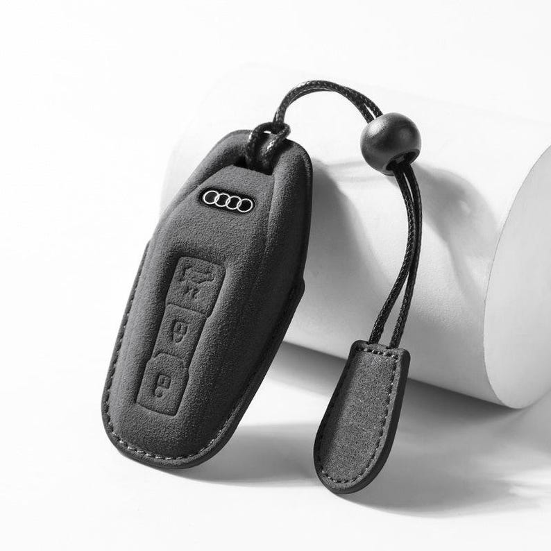 Pode incluir: Uma capa preta para chave de carro com o logotipo da Audi e &iacute;cones de bot&otilde;es. A capa tem um cord&atilde;o com uma conta preta e uma aba de couro. A capa da chave &eacute; feita de um material texturizado.