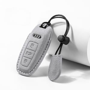 Pode incluir: Uma capa cinza claro para chave de carro com o logotipo da Audi e bot&otilde;es. A capa &eacute; presa a um cord&atilde;o preto com uma pequena etiqueta cinza correspondente. O fundo &eacute; branco neutro.
