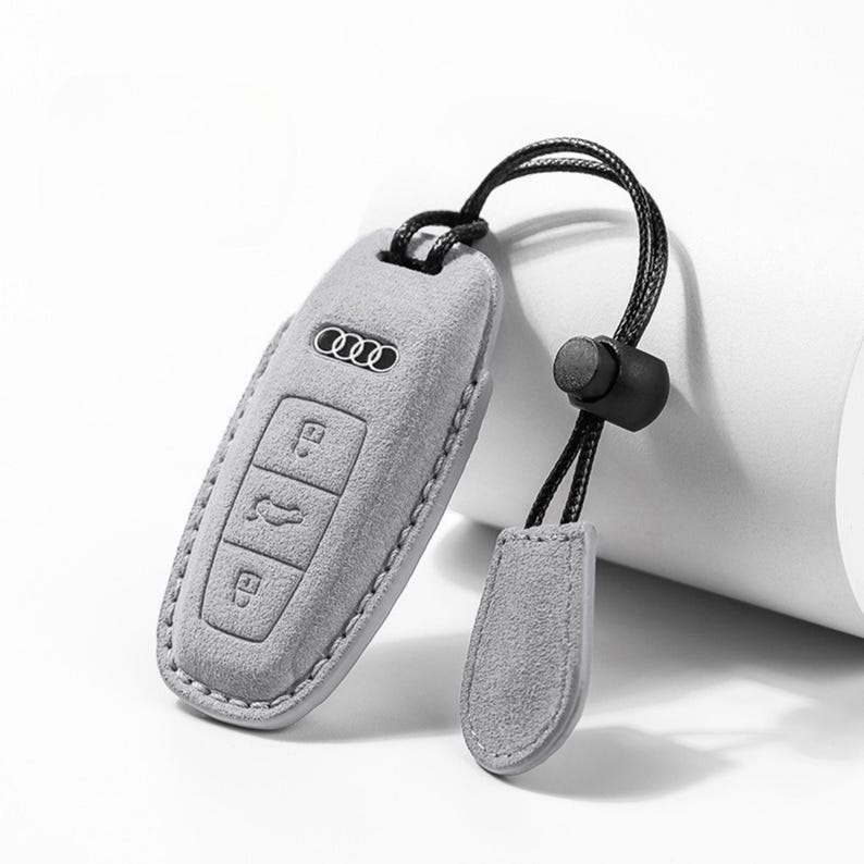 Pode incluir: Uma capa cinza claro para chave de carro com o logotipo da Audi e bot&otilde;es. A capa &eacute; presa a um cord&atilde;o preto com uma pequena etiqueta cinza correspondente. O fundo &eacute; branco neutro.