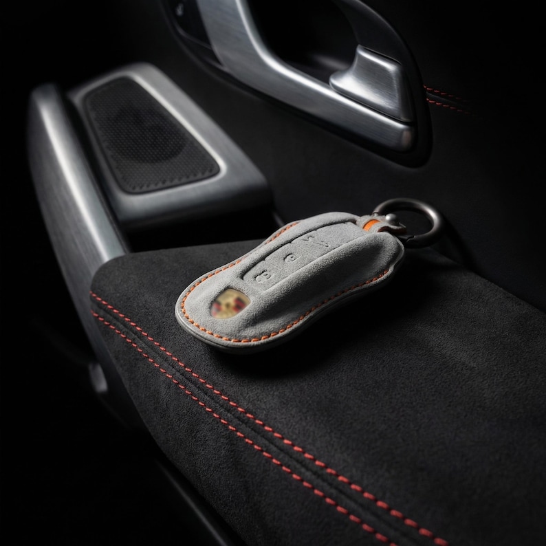 May include: A gray car key fob cover with orange stitching and a metal keyring. The key fob is resting on a black, textured surface with orange stitching. The background includes a car door handle and speaker.