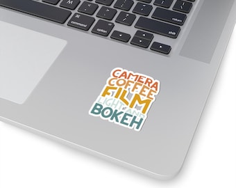 Puede incluir: Una pegatina con las palabras "Camera, Coffee, Film, Light and Bokeh" en un diseño apilado y en capas. Las palabras están en una paleta de colores retro naranja, amarillo y azul.