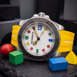 May include: A stainless steel wristwatch with a white face featuring colorful dot hour markers and a date window. The watch has a black bezel and a bright yellow silicone band. The watch face reads "SILVERBACK".