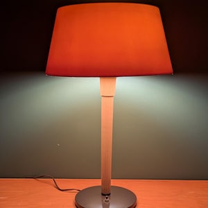 May include: A table lamp with a conical orange lampshade. The lamp has a light-colored wooden stem and a circular, dark gray base. The lamp is plugged into an electrical outlet.