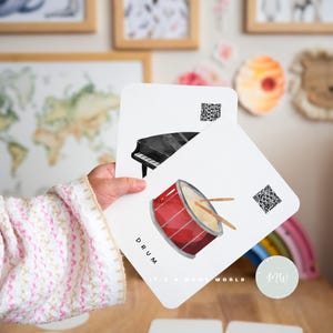 May include: Two flashcards featuring a black and white piano and a red drum with drumsticks. The text "DRUM" is written on the bottom of the card with the drum. The text "IT'S MOMS WORLD" is written on the bottom of the card with the piano.