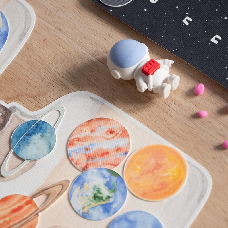 Może przedstawiać: Zestaw kolorowych naklejek z planetami w akwareli z białą gumką do mazania w kształcie astronauty. Planety obejmują niebieską i białą Ziemię, czerwoną i białą Marsa, ż&oacute;łtego i pomarańczowego Jowisza oraz niebieskiego i białego Saturna z pierścieniami.
