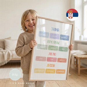 Può includere: Un bambino sorridente tiene in mano un poster incorniciato con testo serbo. Il poster mostra i mesi dell'anno in serbo, ognuno in un colore pastello diverso. La cornice è di colore legno chiaro. La bandiera serba è visibile.