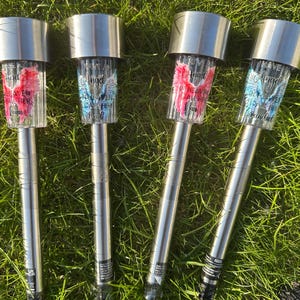 May include: Four solar-powered garden lights with silver tops and stems. The clear plastic light covers display angel wings in pink and blue, with the text "Angel in Heaven". The lights are placed in green grass.