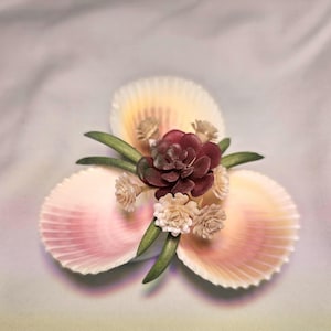 May include: Three seashells arranged in a triangular shape, with a small bouquet of flowers in the center. The flowers are a mix of white and burgundy, with green leaves.