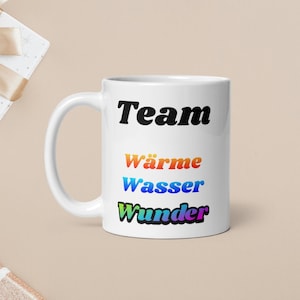 May include: White ceramic mug with the word "Team" in black, and the words "Wärme Wasser Wunder" in rainbow colors. The mug has a white handle and is on a beige surface.