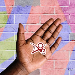 May include: A hand with a red and white sticker on the palm. The sticker features a stylized person with arms raised and a heart shape. The hand is in front of a brick wall with colorful paint.