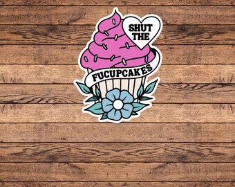 May include: A sticker featuring a pink cupcake with the text "SHUT THE" in a heart and "FUCUPCAKES" on a banner. The cupcake has blue accents and is set against a wooden background.