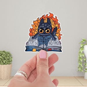 May include: A black cat with flames around its head is holding open a book that says "It's not a slow burn romance if you read fast".
