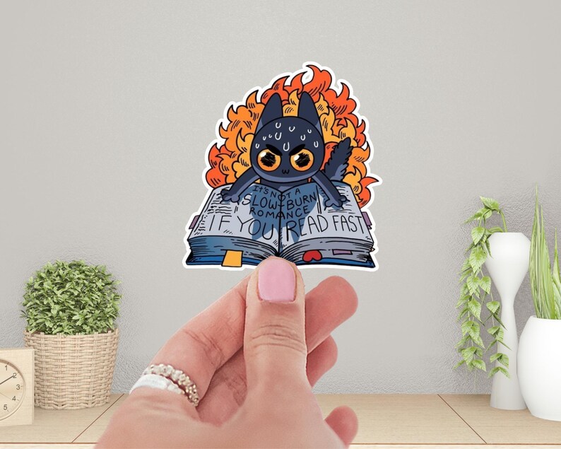 May include: A black cat with flames around its head is holding open a book that says "It's not a slow burn romance if you read fast".