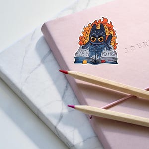 May include: A black cat with orange flames around it is reading a book that says "If you slow burn romance, you read fast".
