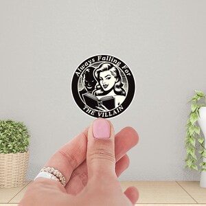 Op de afbeelding: Zwart-witte sticker met een vrouw die een boek leest. De tekst luidt "Always Falling For The Villain".