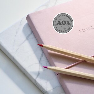 May include: A pink journal with the word "JOURNAL" printed on the cover. A gray sticker with the text "IF THERE IS A SCREEN A03 CAN BE SEEN" is on the cover. Two pencils, one red and one brown, are on top of the journal.