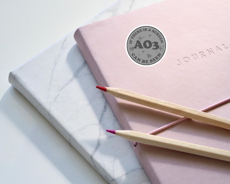 May include: A pink journal with the word "JOURNAL" printed on the cover. A gray sticker with the text "IF THERE IS A SCREEN A03 CAN BE SEEN" is on the cover. Two pencils, one red and one brown, are on top of the journal.