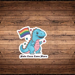 Pode incluir: Um dinossauro azul de desenho animado segurando uma bandeira arco-íris do orgulho com o texto "Hate Less Love More".