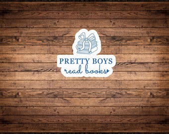 Könnte beinhalten: Ein Aufkleber mit einem Stapel Bücher, die mit einer Schleife gebunden sind, mit dem Text "PRETTY BOYS read books" in Blau. Der Aufkleber befindet sich auf einem Holzgrund.