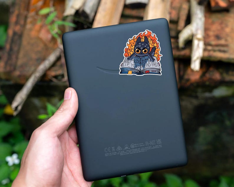 May include: A black Kindle e-reader with a sticker of a black dragon reading a book with the text "Read fast if you want to live".