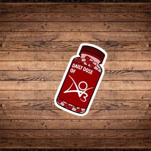 May include: A red and white sticker with a pill bottle design. The bottle is filled with pink pills and has the text "Daily Dose of" and a logo with the letters "V3".