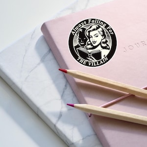 Op de afbeelding: Een zwart-witte sticker met de tekst "Always Falling For The Villain" en een illustratie van een vrouw die een boek leest met een man achter haar.