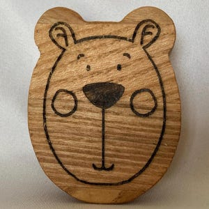 May include: A wooden bear face ornament with a simple, hand-drawn design. The bear has a black nose, black eyes, and a small smile.