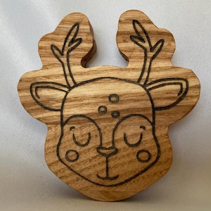 以下が含まれることがあります： 木製のトナカイのオーナメント。茶色の木目調仕上げで、黒い線で描かれたトナカイの顔に、角、目、鼻が描かれています。
