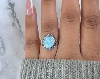May include: A silver ring featuring a light blue oval gemstone, set within a decorative, openwork frame. The ring is displayed on a hand wearing a gray sweater, with manicured nails in the background. The gemstone has a marbled appearance.