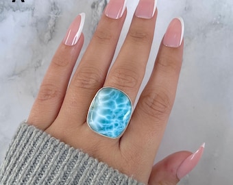 May include: A silver ring featuring a large, square-shaped larimar stone. The stone has a light blue color with white marbling, set in a polished silver band. The ring is displayed on a hand with long, manicured nails.