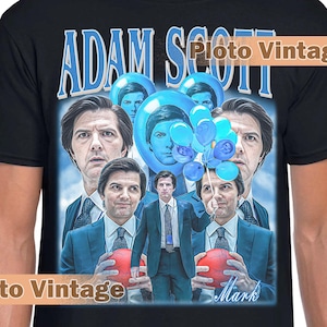 May include: Black t-shirt featuring a graphic of Adam Scott with a blue and white design. The design includes the text "ADAM SCOTT" in blue, along with multiple images of the actor. The shirt has a crew neck.