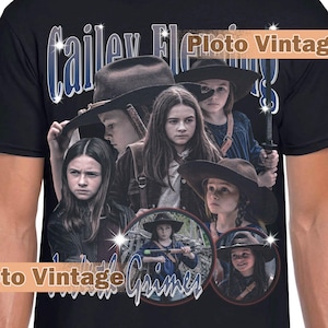 May include: Black t-shirt featuring a collage of images with the text "Cailey Fleming Grimes" and "Ploto Vintage." The design includes images of a young girl wearing a hat and holding a weapon.