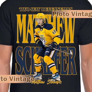 May include: Black t-shirt featuring a hockey player in a yellow and navy uniform. The shirt displays the name "Matthew Schaefer" in large, stylized lettering, along with the text "Two-Way Defenseman."