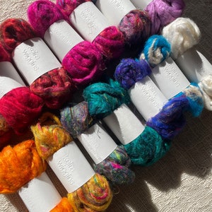 May include: A collection of colourful, hand-dyed fibre rovings, each labelled with "Koira Wiggins". The rovings are arranged in a fan shape, with the colours ranging from purple to orange.