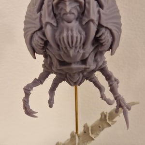 May include: A detailed, gray miniature figure of a creature with a large head, multiple limbs, and a textured body. The figure is mounted on a base with a gold rod. The overall design suggests a science fiction or fantasy theme.