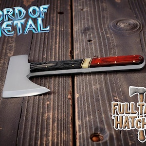 May include: A full tang hatchet with a silver blade and a handle featuring black, cream, and red wood. The text "LORD OF METAL" and "FULL TANG HATCHET" are visible. The hatchet is displayed on a wooden surface.