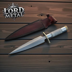 May include: A silver dagger with a pointed blade and a textured handle, resting on a wooden surface. The dagger has a gold crossguard and is accompanied by a brown leather sheath. The logo "LORD of METAL" is visible in the upper left corner.