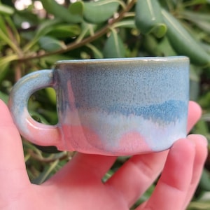 Puede incluir: Una pequeña taza de cerámica hecha a mano con un asa redondeada. La taza presenta un degradado de colores, incluyendo azul, blanco y rosa. La taza se sostiene en una mano, con un fondo verde borroso.