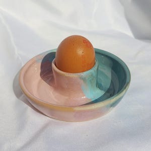 Puede incluir: Un huevera de cerámica con una base redonda y poco profunda con un soporte central. El esmalte muestra una paleta de colores mezclados de rosa pastel suave, azul claro y toques de amarillo. Un huevo de gallina marrón descansa en el centro.
