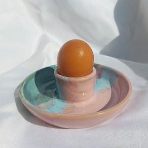 Puede incluir: Un huevera de cerámica con una base circular ancha y poco profunda y un soporte cilíndrico central. El esmalte muestra un patrón de acuarela mezclado en tonos rosa pastel suave, azul claro y toques de amarillo. Un huevo marrón descansa dentro.